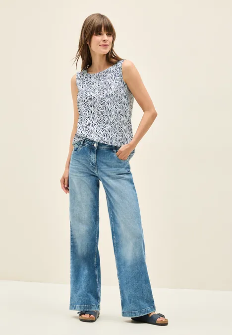 Frau in weitem Jeans und gemustertem Top, lässig stehend, helle Umgebung.